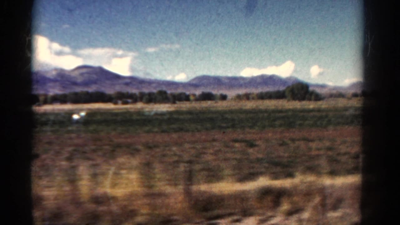 Moving train view across open fields with mountains, clouds, and a small white vehicle in the distance.