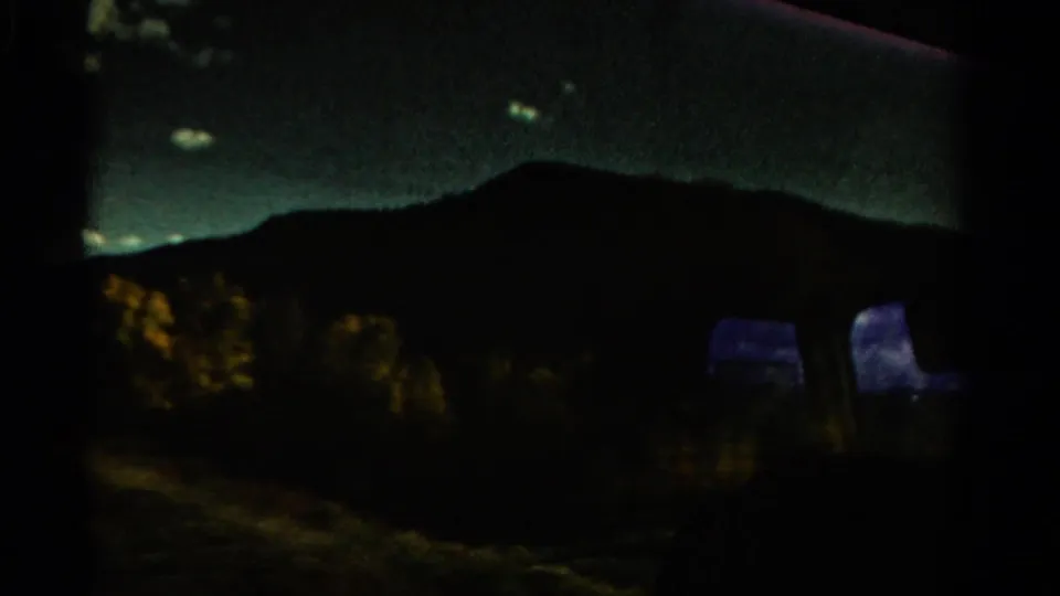 A darker Colorado travel view from the train shows a mountain silhouette and shadowed vegetation, with glass reflections suggesting changing light.
