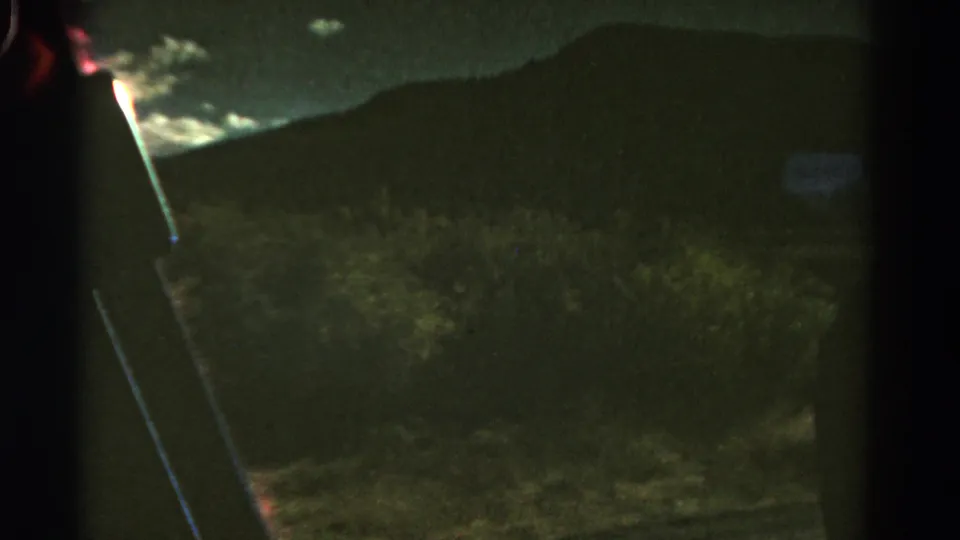 1967 Colorado moving-car frame showing silhouetted mountains, roadside brush, a bright cloud patch, and a faint pale shape.