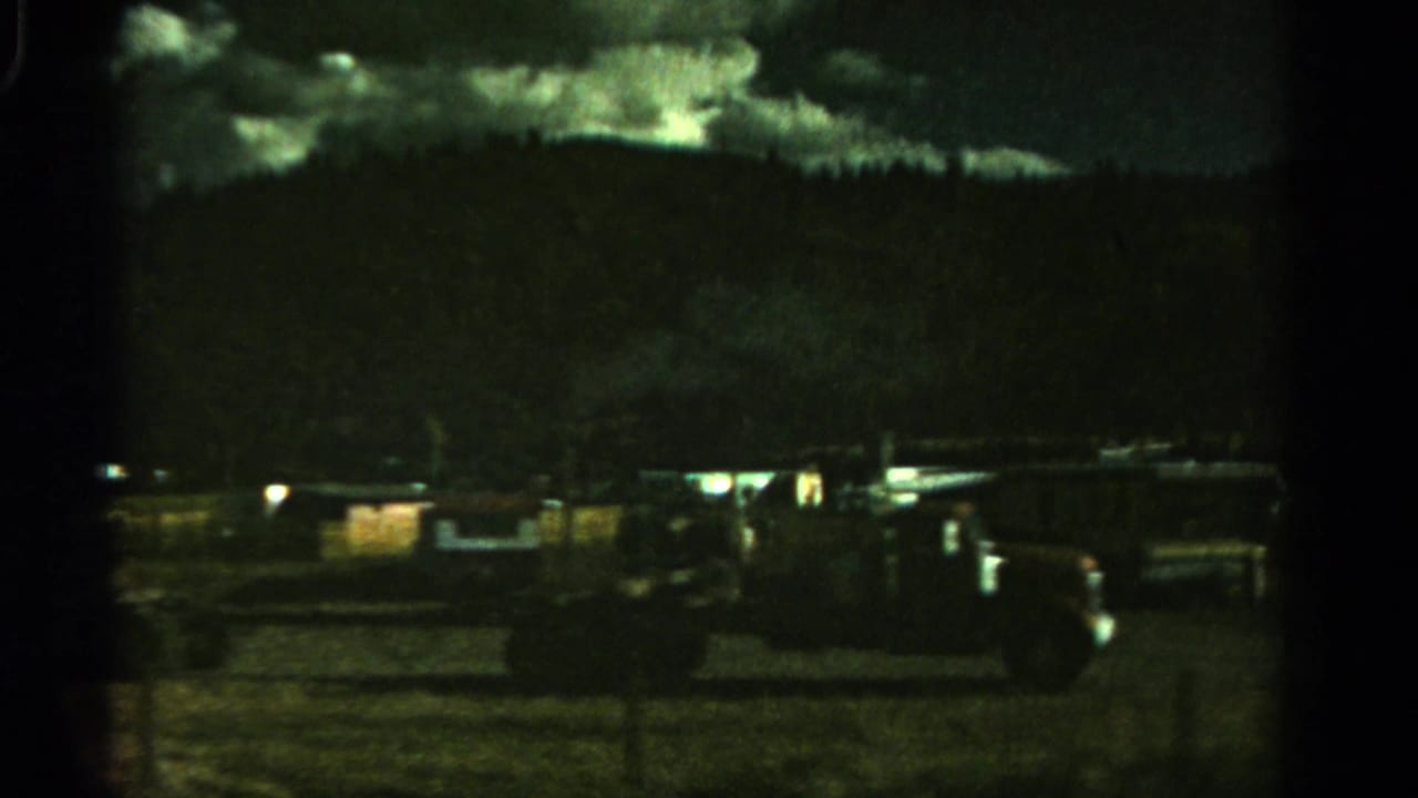 Dark trailer park view with mobile homes, parked vehicles, hills, and cloudy sky.