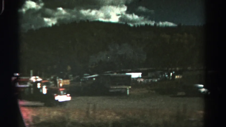 Wide 1967 Colorado home-movie view of a dim trailer park with mobile homes, parked cars, open ground, and heavy clouds.