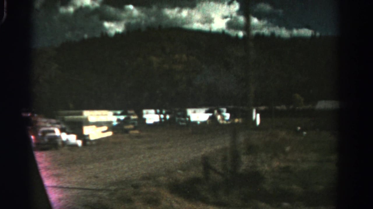Moving view past cars, bright camper lights, a gravel area, trees, and a mountain slope.