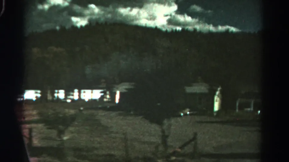 1967 Colorado home-movie frame with campers and parked cars near posts in a dim gravel lot below a mountain ridge.