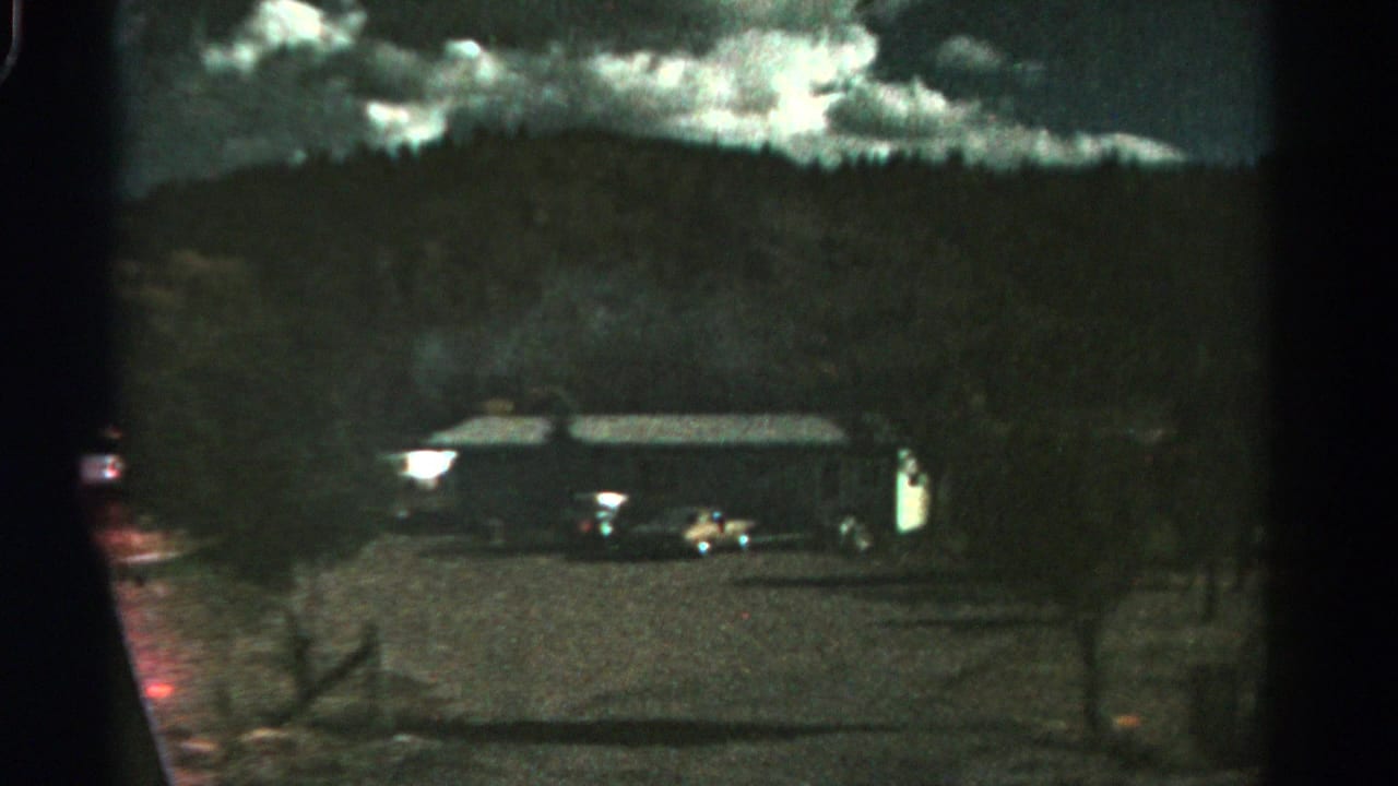 Dark rural building below a wooded hillside with parked cars and a cloudy sky.