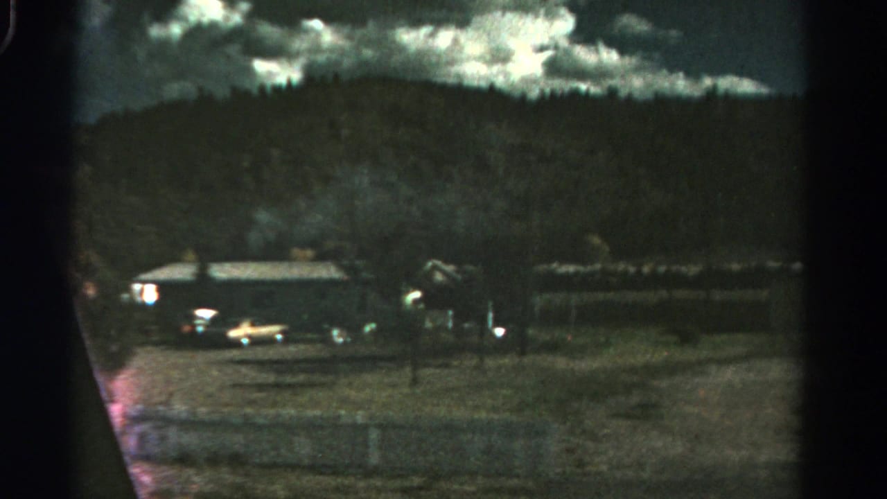 Dimly lit rural yard with parked cars, a long building, fence posts, and hillside.