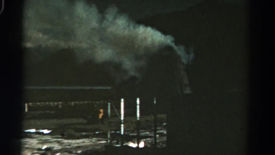 1967 Colorado 8mm frame with a pale smoke cloud drifting over dark industrial structures and bright reflections near the ground.