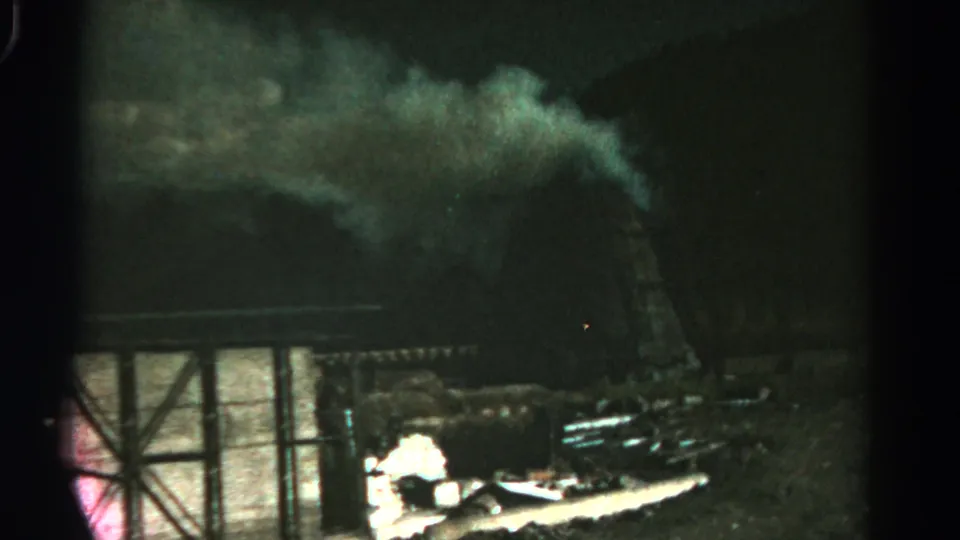 Nighttime 8mm view in Colorado, 1967, with smoke over a dark structure and bright firelight beside scattered material.