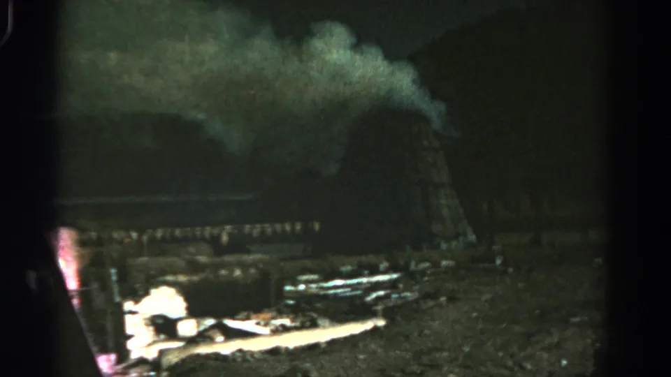 Grainy 1967 Colorado 8mm frame of a nighttime fire scene, with smoke, wooden framing, and a bright glow near ground debris.