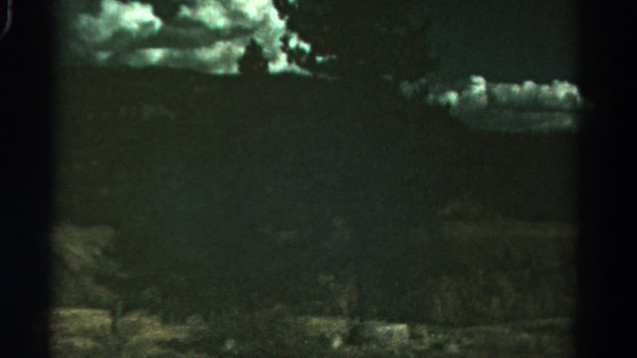 1967 8mm Colorado home-movie frame showing shadowed hills, a tree line, open foreground terrain, and bright textured clouds.
