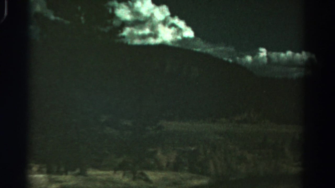1967 Colorado landscape frame with a broad mountain ridge, glowing clouds, trees, and shadowed foreground vegetation on aged 8mm film.