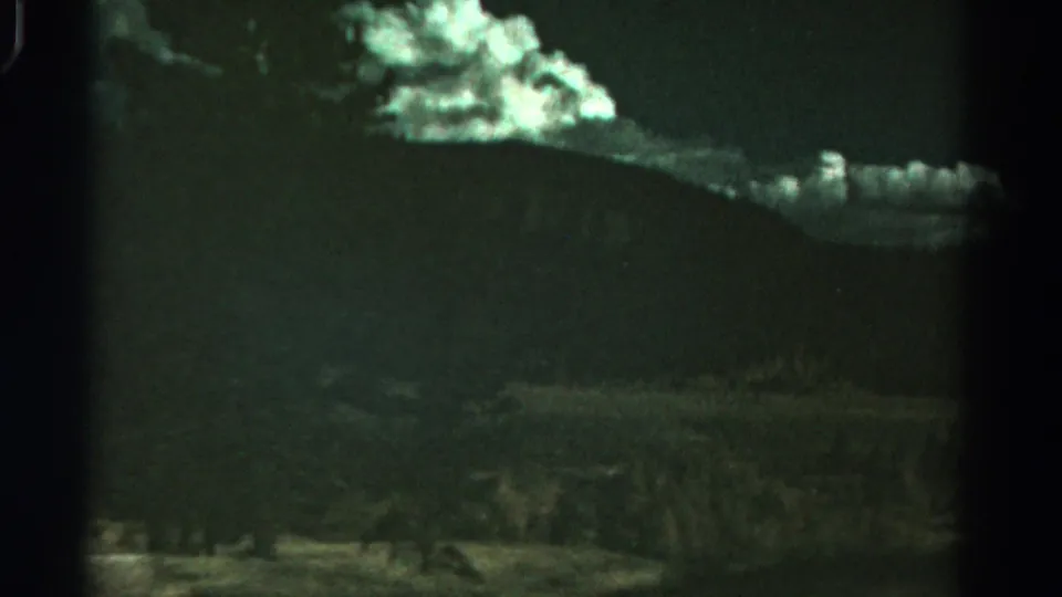 1967 Colorado landscape frame with a broad mountain ridge, glowing clouds, trees, and shadowed foreground vegetation on aged 8mm film.