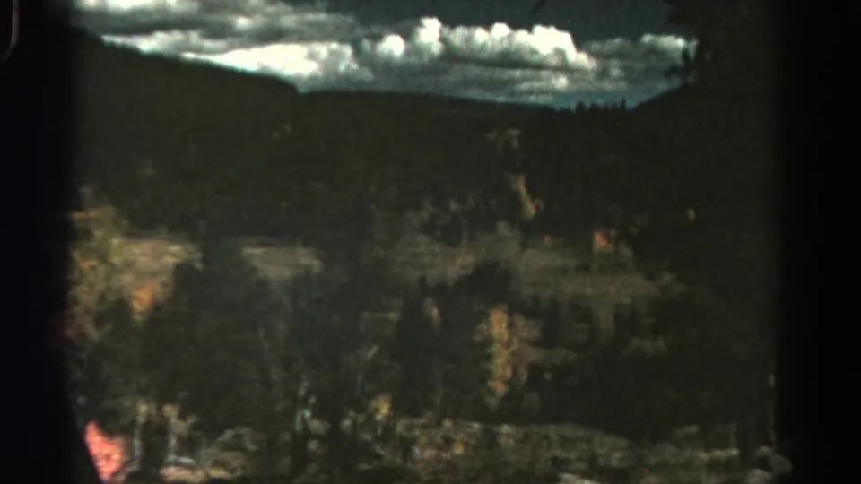 1967 Colorado countryside passes by through a car window, with clouds, trees, hills, and film-edge darkness.
