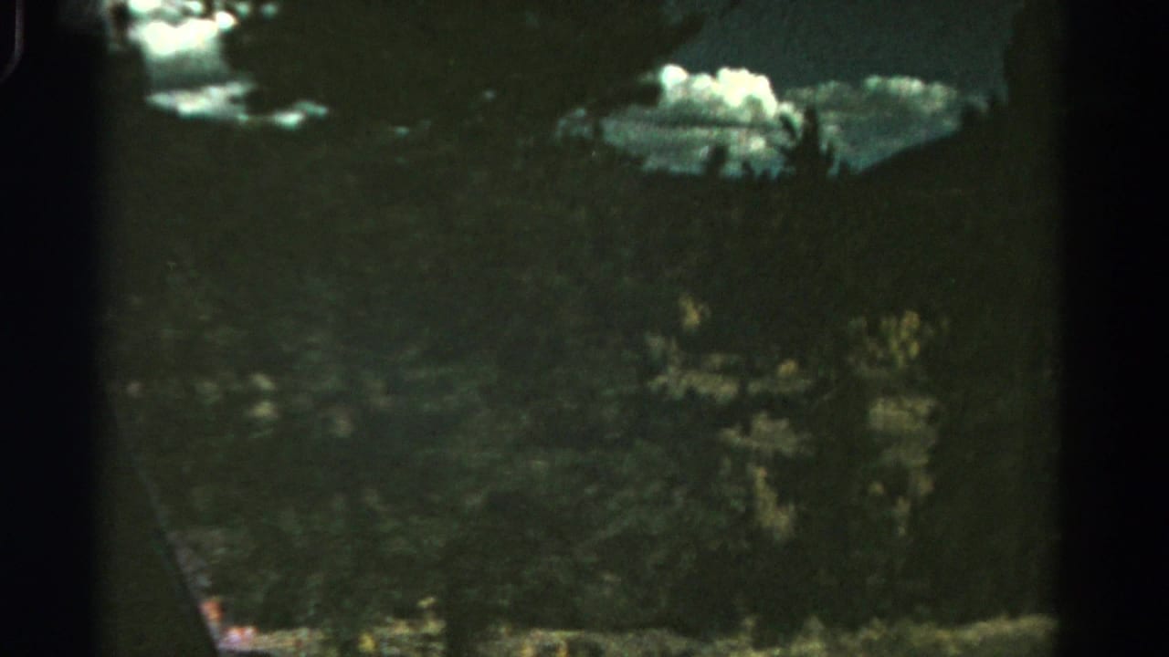 1967 Colorado 8mm frame showing a dark evergreen-covered hillside below bright cloud patches and soft mountain forms.