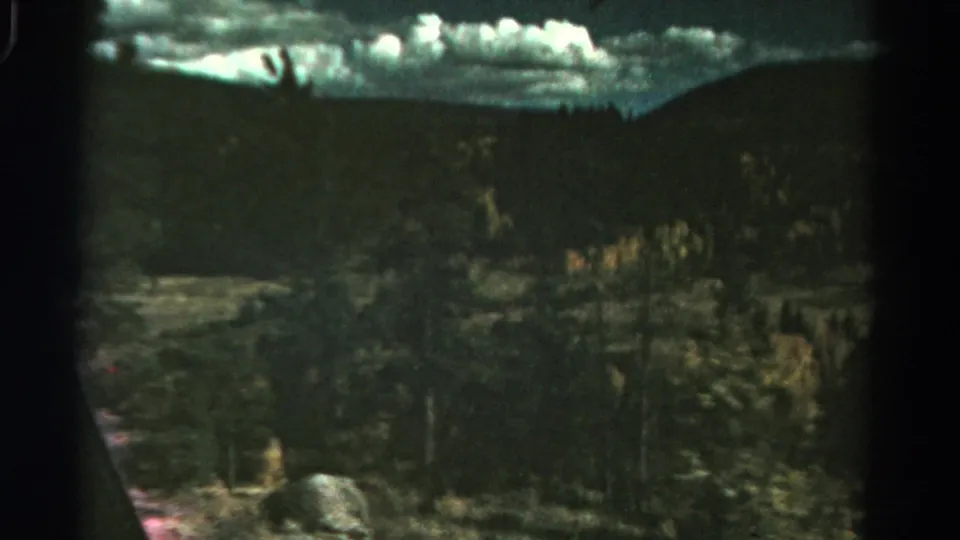 1967 Colorado 8mm frame showing evergreen trees, a pale grassy clearing, and clouds above a distant hillside.