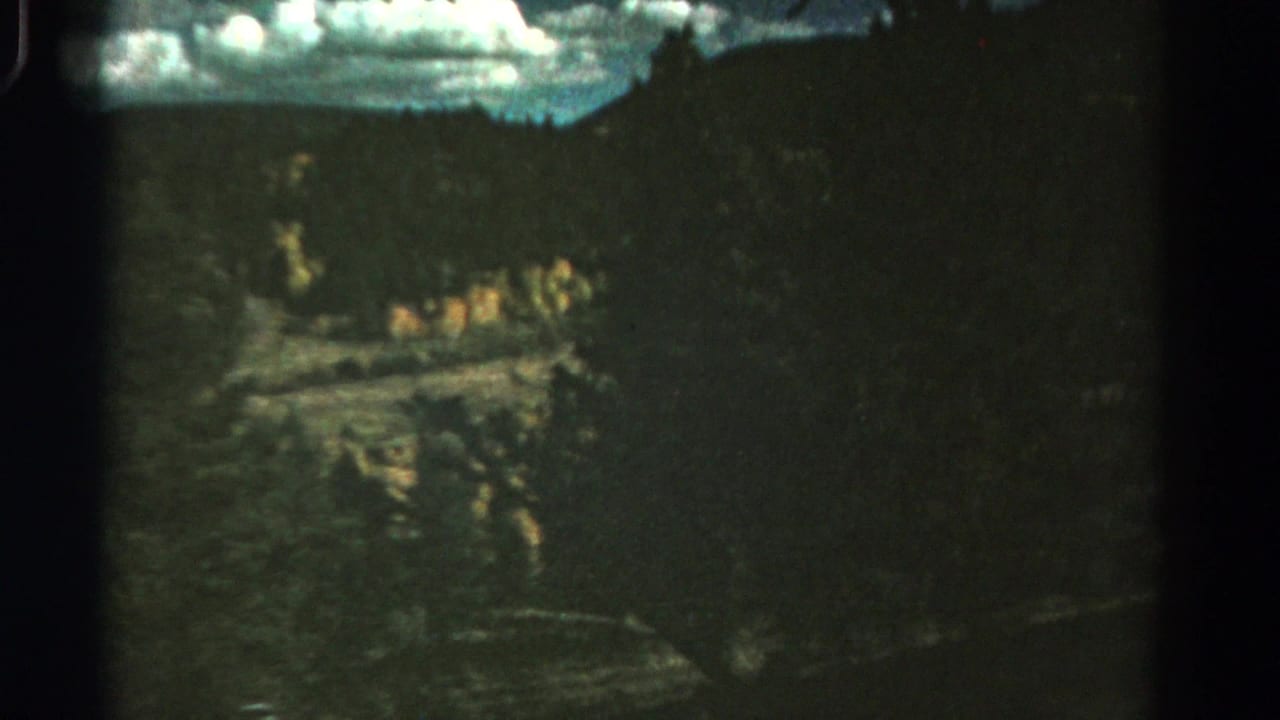 1967 Colorado 8mm frame showing a shadowed wooded roadside drop with cloudy sky and blurred terrain from a moving vehicle.