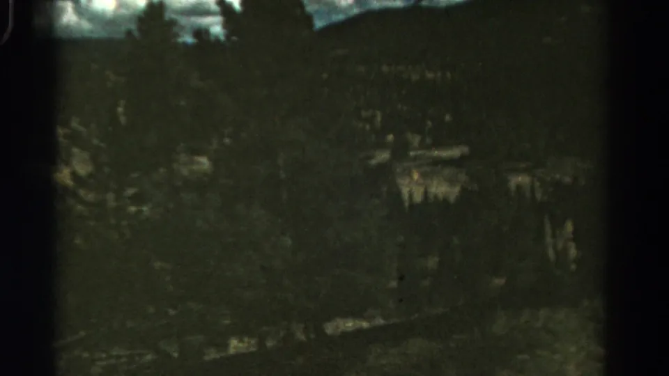 1967 Colorado travel footage frame with dense trees, a steep drop, and dark green color shift visible through 8mm film grain.