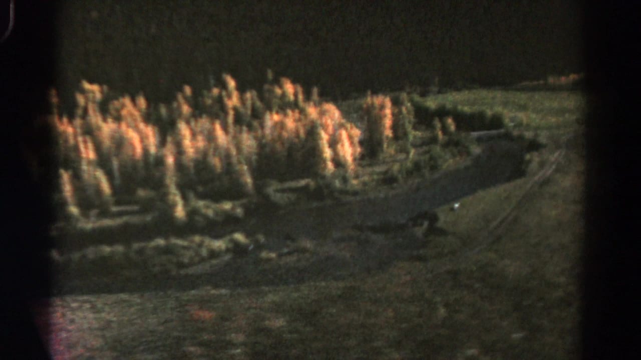 1967 Colorado 8mm frame showing a shadowed valley, warm tree clusters, and a pale winding path or stream from a high viewpoint.