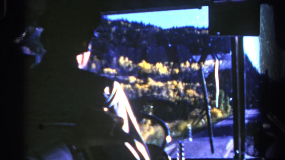 Inside a 1967 Colorado bus, a silhouetted driver is seen near the windshield with autumn trees and hills outside.