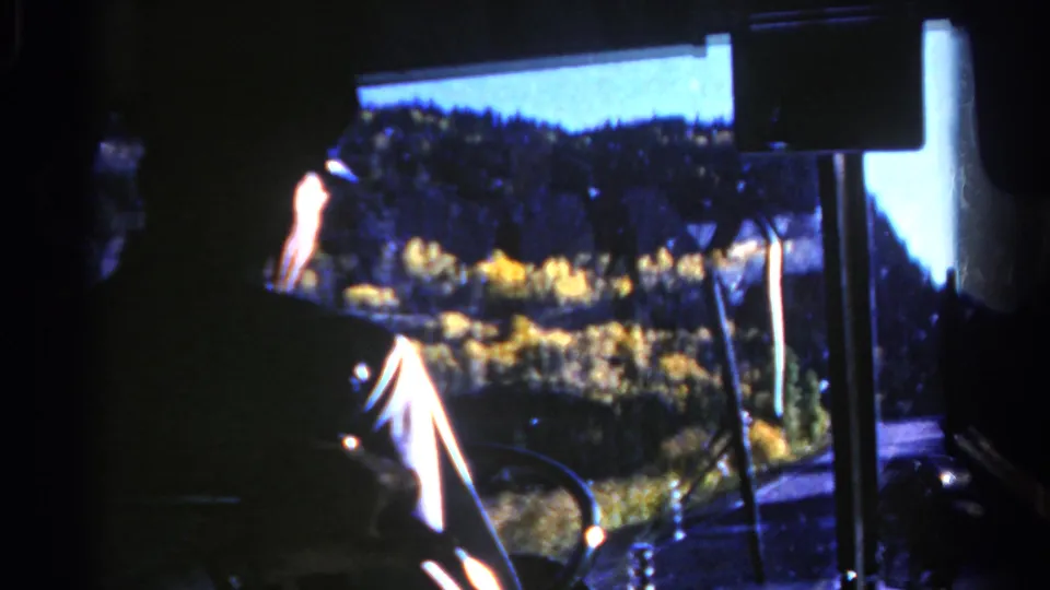 A 1967 Colorado bus interior view shows the driver in silhouette against golden trees and forested hills through the windshield.