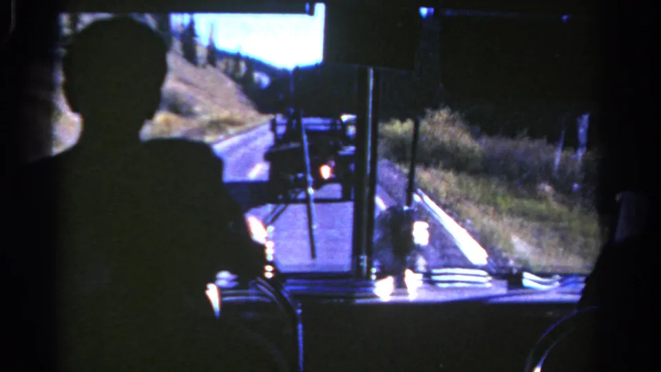 Interior bus view in Colorado, 1967, with a dark passenger silhouette, windshield posts, and a truck ahead on a curving road.