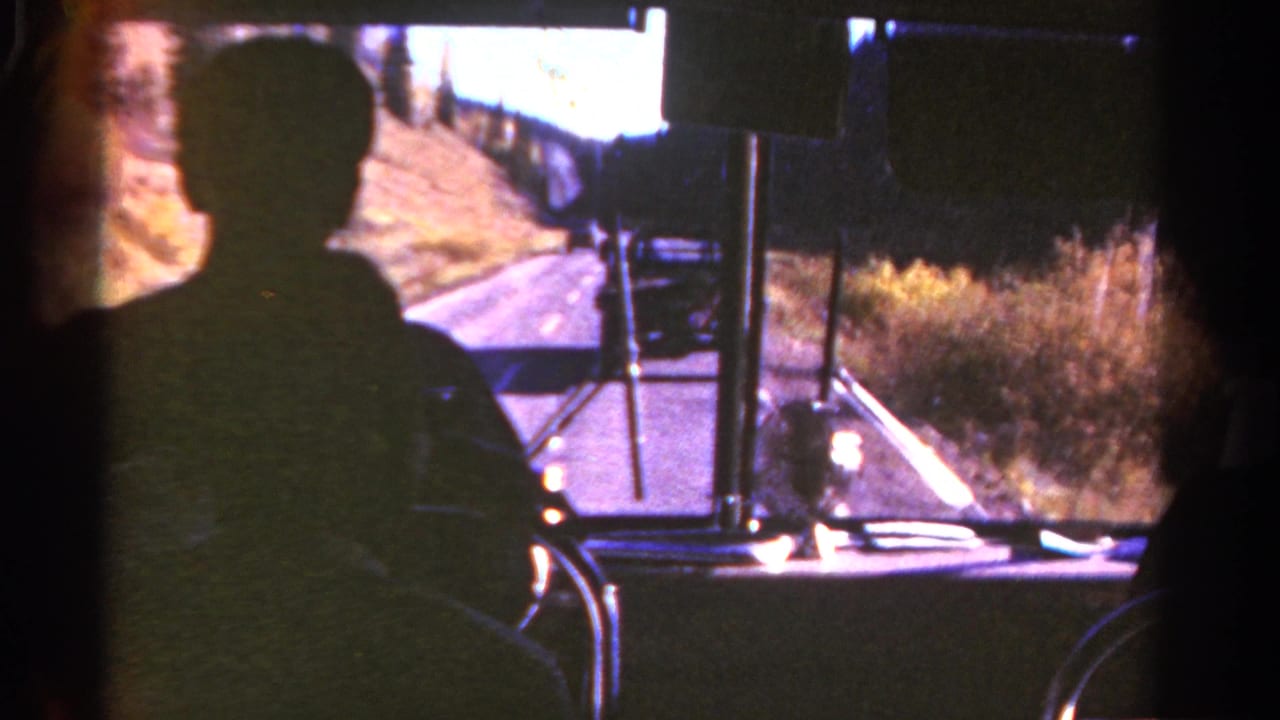 Driver silhouette and bus windshield with a road curving past dry hillside vegetation.