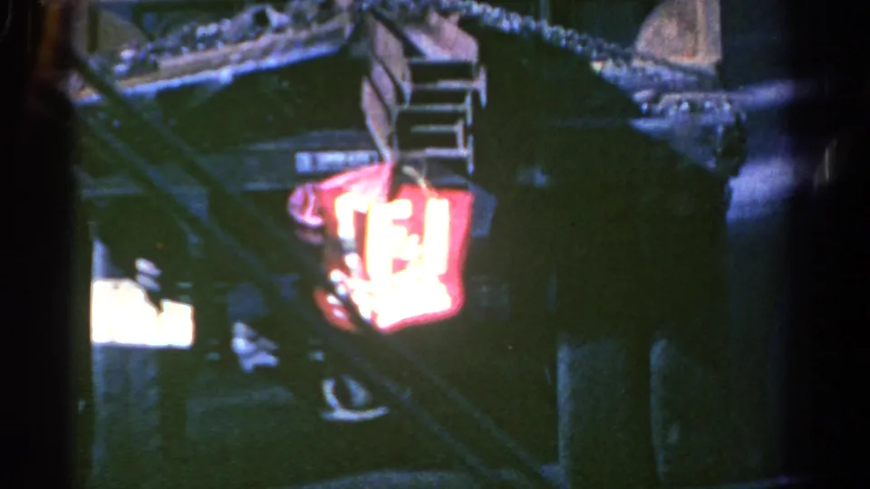 1967 8mm bus interior frame with passengers in low light near handrails and a bright red illuminated sign or light.