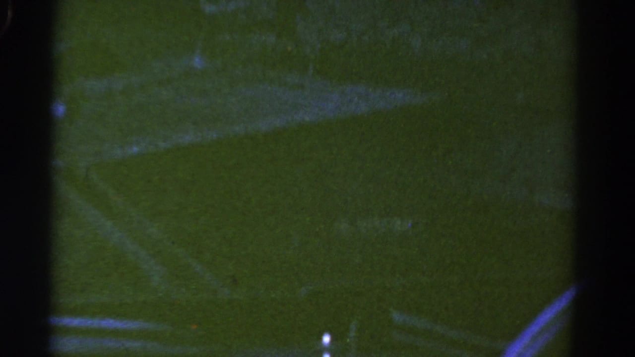 1967 Colorado home-movie frame looking through a dark car window at faint roadway markings and green-tinted roadside ground.