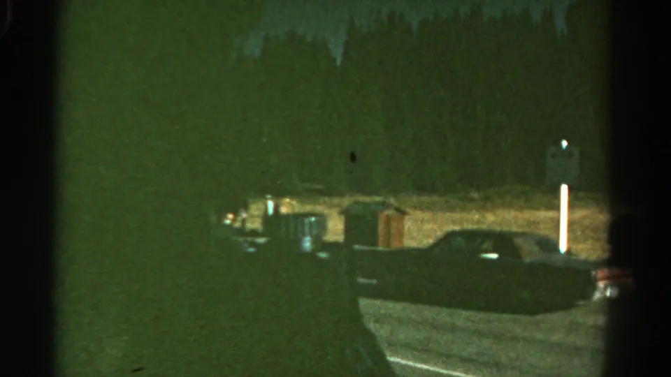 1967 Colorado roadside frame with a dark parked car shape, vertical roadside posts, and grainy low-light 8mm texture.