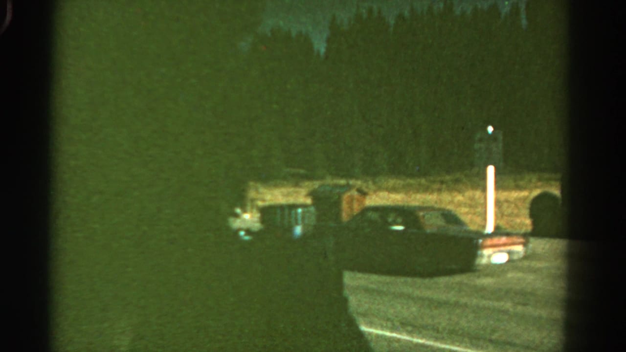 1967 Colorado home-movie frame showing a car ahead on a dark road, with roadside posts and forest darkness lit by vehicle headlights.