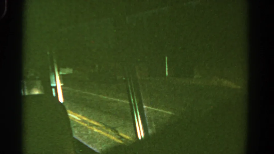 1967 8mm frame from inside a moving vehicle, showing a window edge, yellow road lines, and a dark wooded roadside in low light.