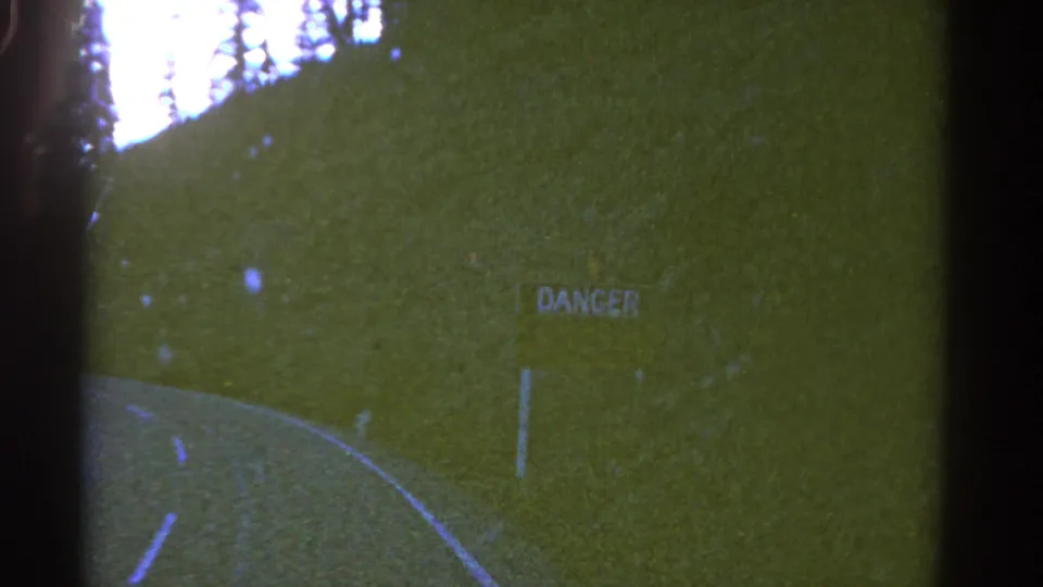 1967 Colorado road-travel frame showing a curving paved road, wooded hillside, and DANGER sign in grainy 8mm color.