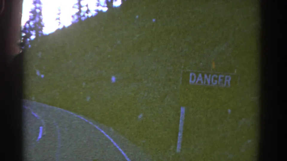 1967 Colorado home-movie frame with a closer roadside DANGER sign, grassy slope, road curve, and soft 8mm grain.