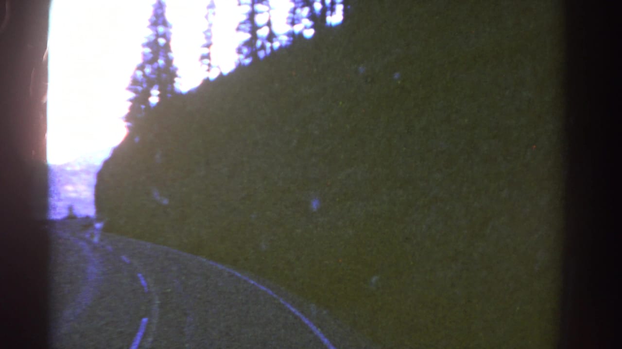 1967 Colorado 8mm frame showing a curving road through shadowed trees with mountain ridges beyond.