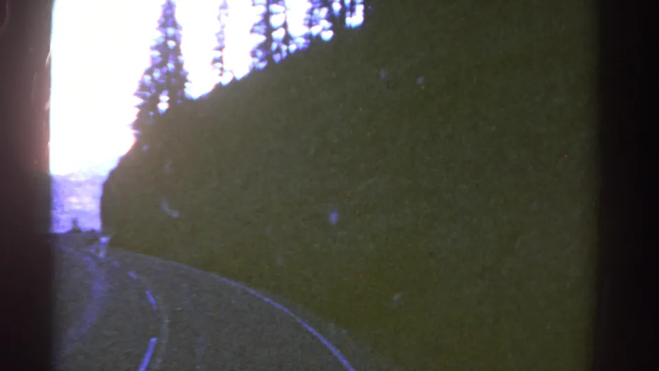 1967 Colorado 8mm frame showing a curving road through shadowed trees with mountain ridges beyond.