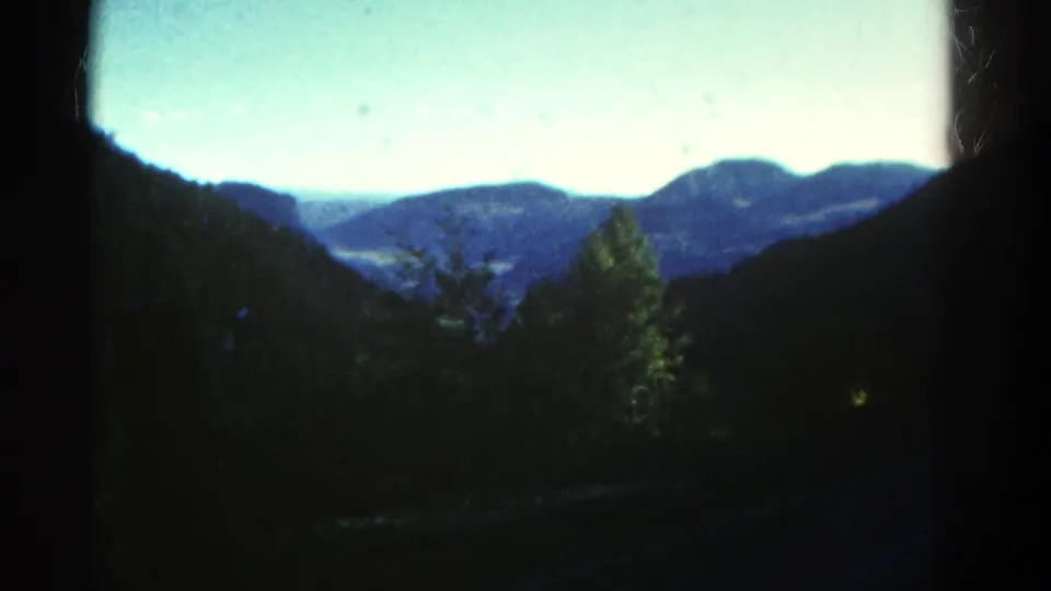 1967 Colorado home-movie frame of layered mountain ridges, roadside trees, and dark window framing from a moving vehicle.