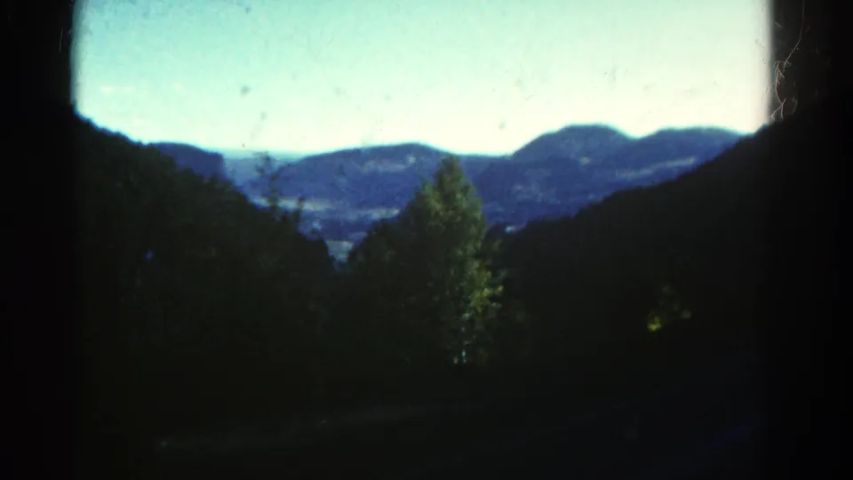 1967 Colorado 8mm frame showing a dark-edged view across foreground trees toward a valley and blue mountain ridges.