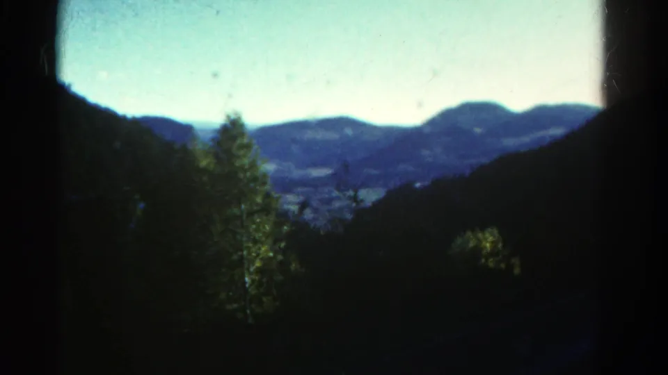 1967 Colorado home-movie frame looking past trees and a dark frame edge toward a valley and rounded mountain peaks.