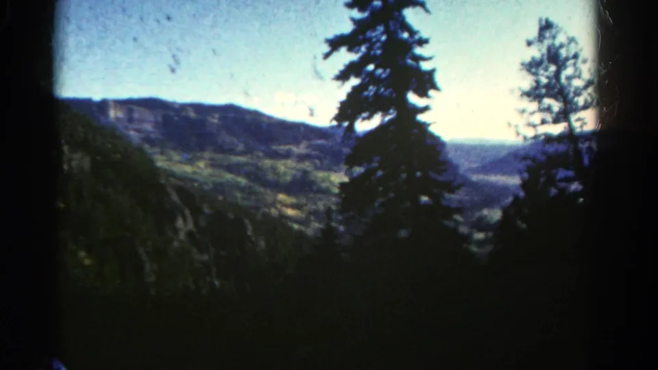 1967 Colorado frame with evergreen silhouettes, rolling mountain hills, and bright sky in vintage 8mm color.