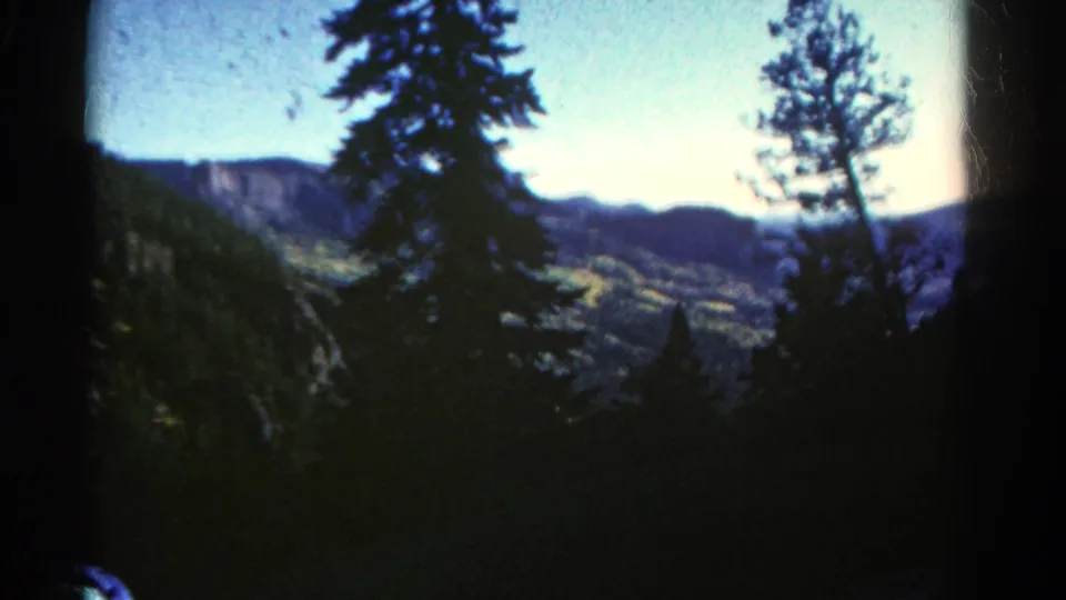 Colorado 8mm travel footage frame with roadside trees, a bright green valley, and distant ridgelines under blue sky.