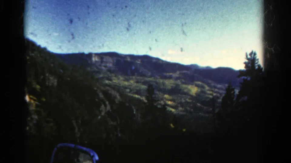 Final 1967 Colorado frame from the moving-car view, looking across dark trees toward a green valley and layered ridges.