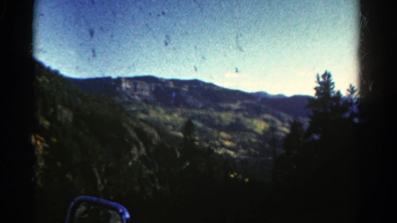 1967 Colorado 8mm frame showing distant cliffs, forested slopes, blue sky, and a car side mirror from a roadside passenger view.