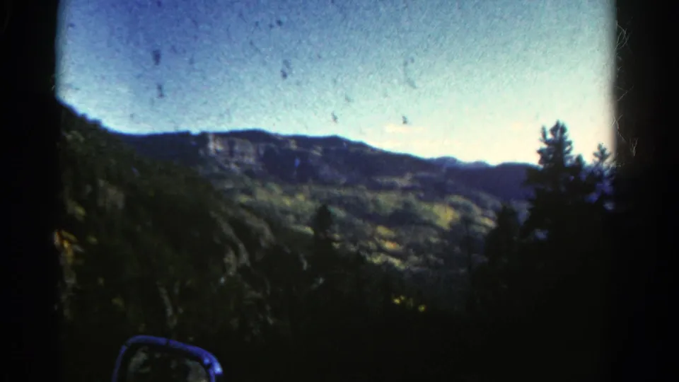1967 Colorado 8mm frame showing distant cliffs, forested slopes, blue sky, and a car side mirror from a roadside passenger view.