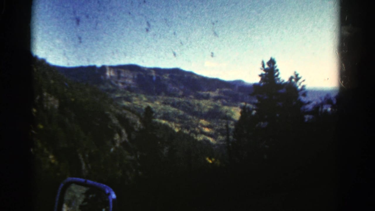 1967 Colorado travel frame with dark roadside trees, distant cliff formations, valley terrain, and a car side mirror below.