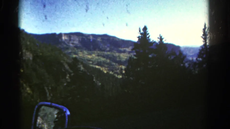 1967 Colorado 8mm view of cliffs, green valley slopes, evergreen silhouettes, and a side mirror during a mountain drive.