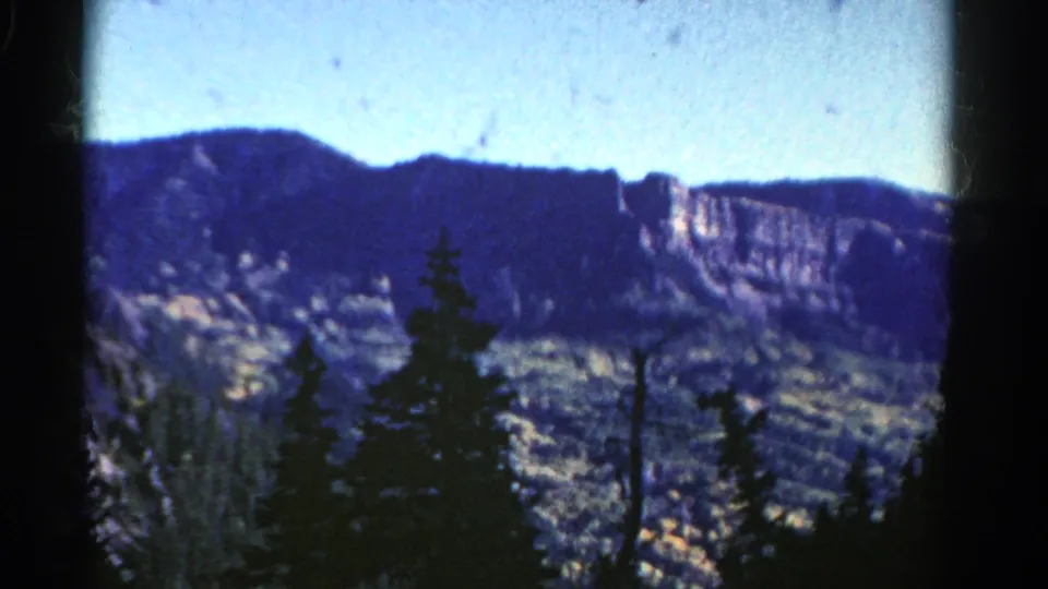 1967 Colorado 8mm frame with a closer cliff wall, dark ridge, scattered evergreens, and hazy valley terrain.