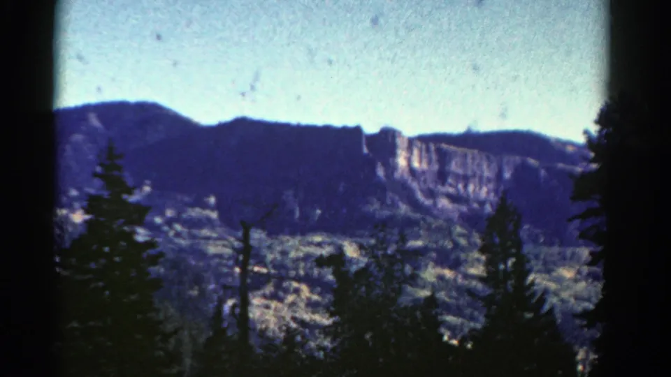 1967 Colorado 8mm view of distant cliffs and slopes partially obscured by dark foreground evergreens.