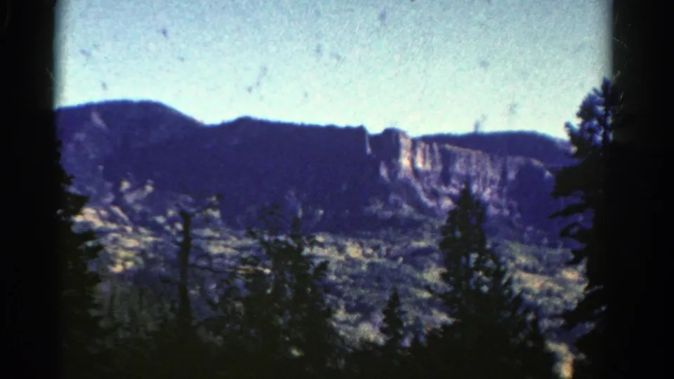 1967 Colorado home-movie frame showing rocky cliffs and wooded hills beyond silhouetted evergreen trees.