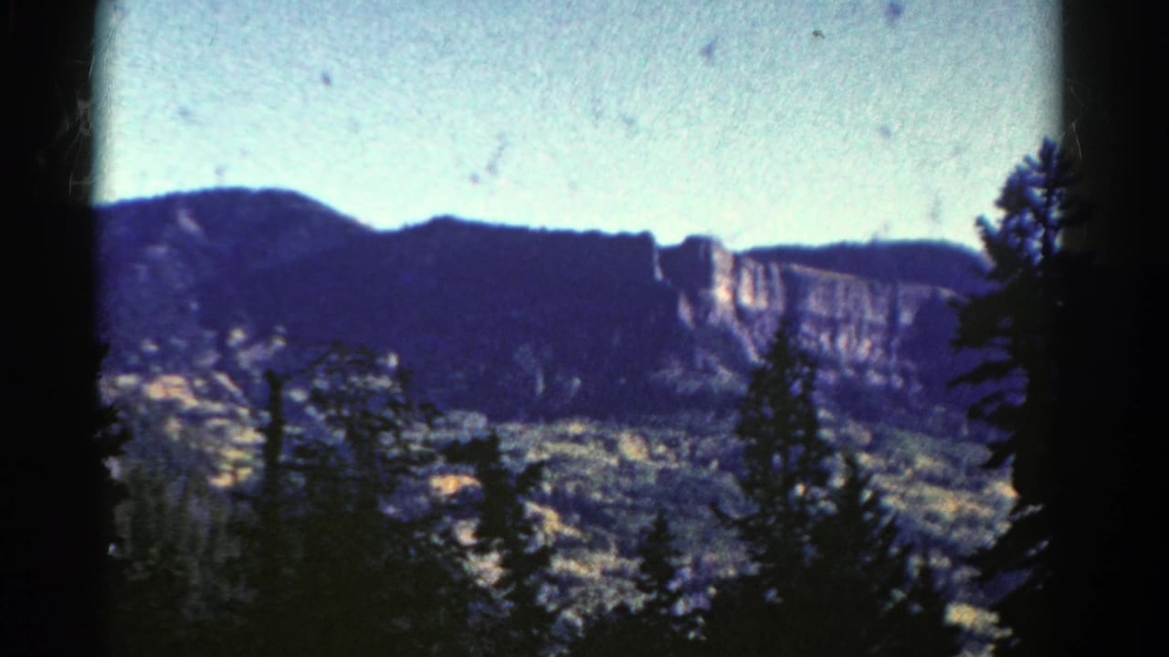 1967 Colorado 8mm frame showing dark evergreen silhouettes, a broad wooded valley, and a sunlit rocky cliff below a pale sky.