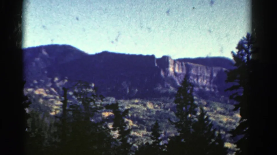 1967 Colorado 8mm frame showing dark evergreen silhouettes, a broad wooded valley, and a sunlit rocky cliff below a pale sky.