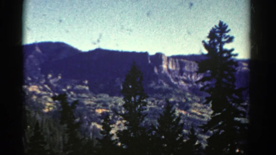 1967 Colorado 8mm frame with foreground conifers, a forested valley, distant rocky cliff, and visible film grain and specks.